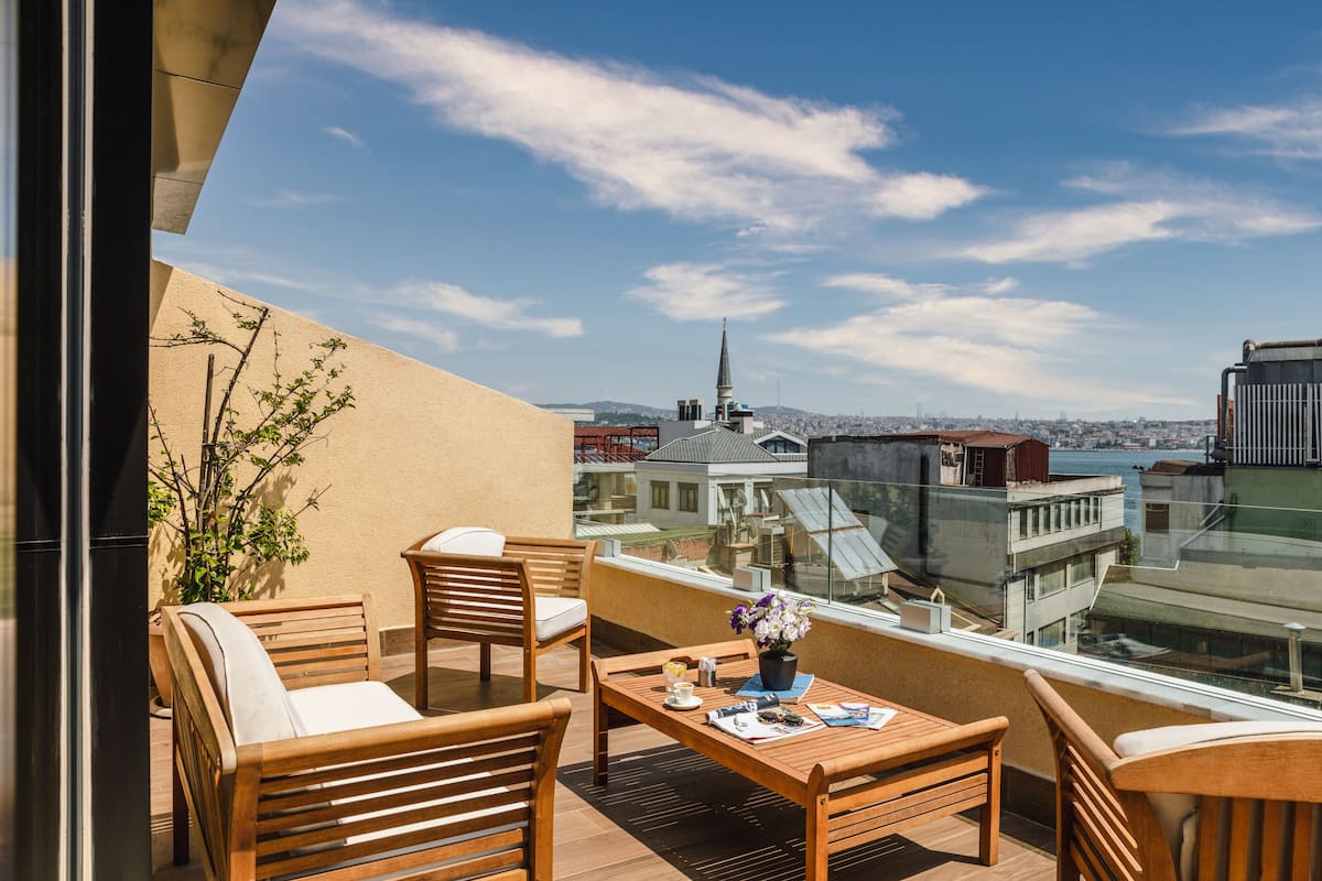 Ein Balkon mit Holzmöbeln, einem Tisch mit Blumenvase und Blick auf eine Stadtlandschaft mit Gebäuden und einem Turm.