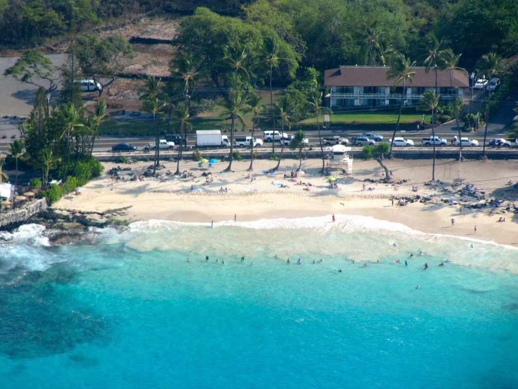 An aerial view of a beach with clear turquoise waters, palm trees, and a building in the background.