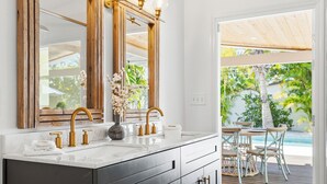 Une salle de bain dotée d’un grand miroir, de robinets dorés et d’un vase rempli de fleurs posé sur le meuble-lavabo.