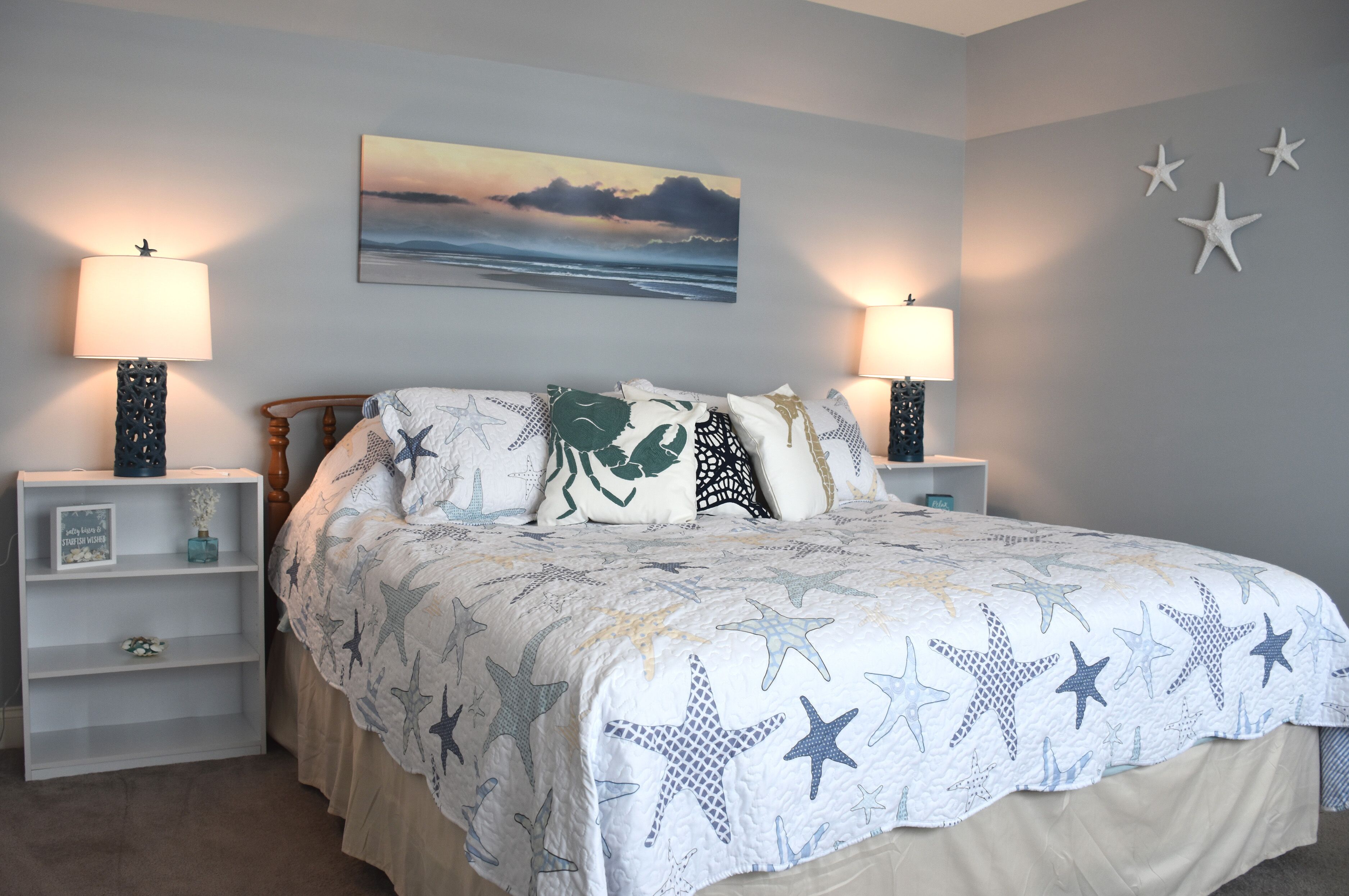 A bedroom with a bed, two bedside tables, a nightstand, a wall-mounted lamp, and a painting of a sunset over the ocean.