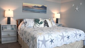 Een slaapkamer met een bed, twee nachtkastjes, een wandlamp en een schilderij van een zonsondergang boven de oceaan.