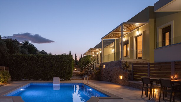 Un área de piscina moderna con una piscina azul cristalina, escalones de piedra que conducen a un espacio exterior cubierto, y un conjunto de mesas y sillas.