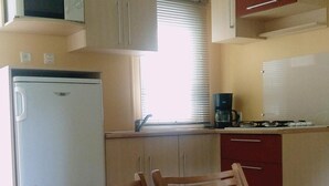 A compact kitchen with a refrigerator, microwave, and coffee maker. There is a dining table with chairs and a window with blinds. - Mobil Home 6 Personnes, 3 Chambres et Terrasse Couverte (Boofzheim)