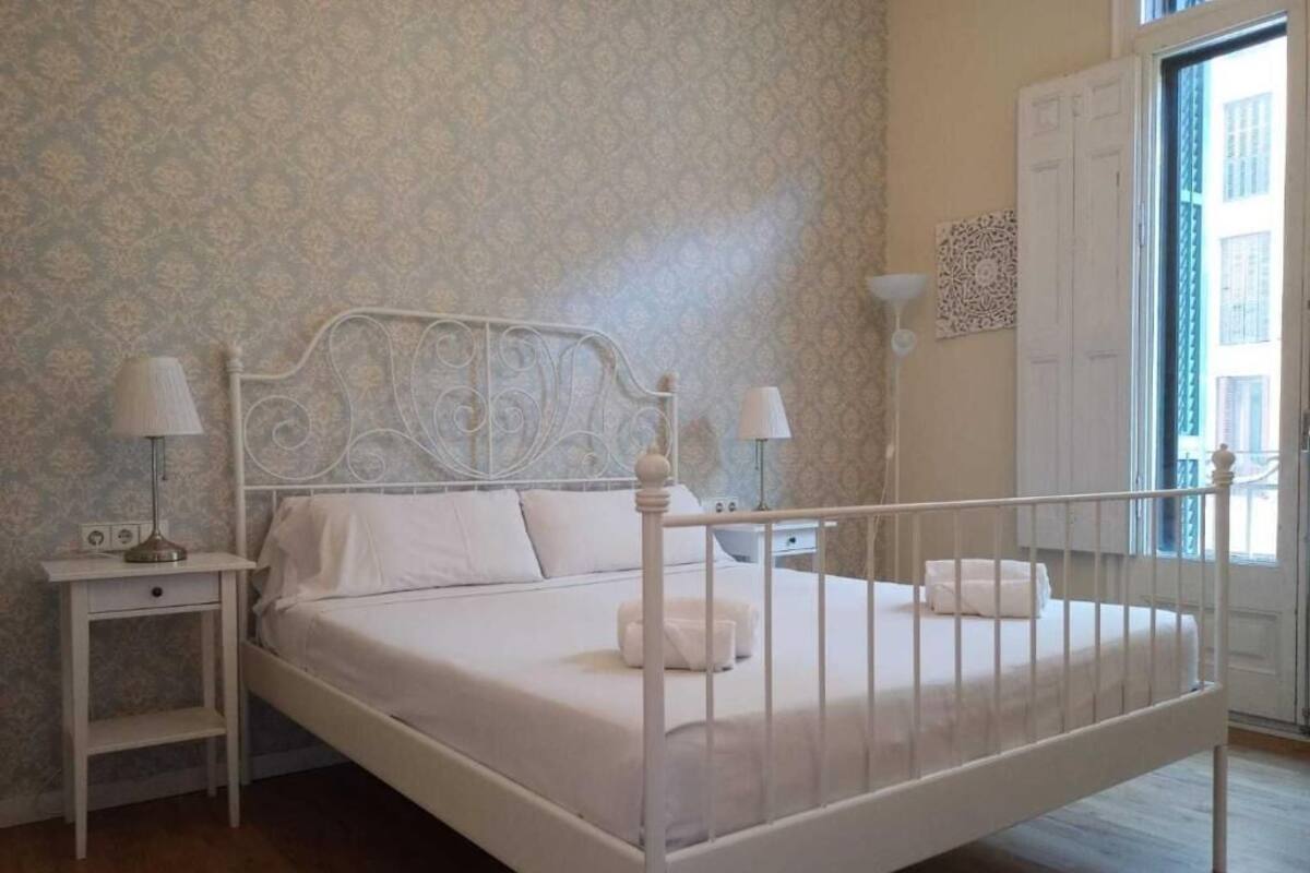 A neatly made bed with white linens and a metal headboard, two bedside tables with lamps, and a patterned wallpaper.