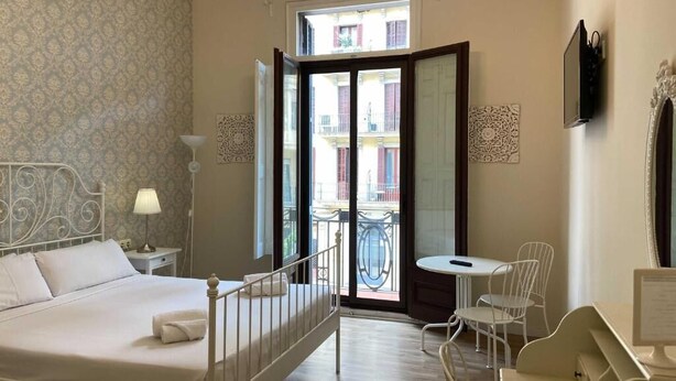 Hotelkamer met een bed, bureau, stoel, klein tafeltje, lamp, televisie en een balkon met uitzicht op gebouwen.