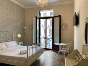 Ein Hotelzimmer mit Bett, Schreibtisch, Stuhl, kleinem Tisch, Lampe, Fernseher und einem Balkon mit Blick auf Gebäude.