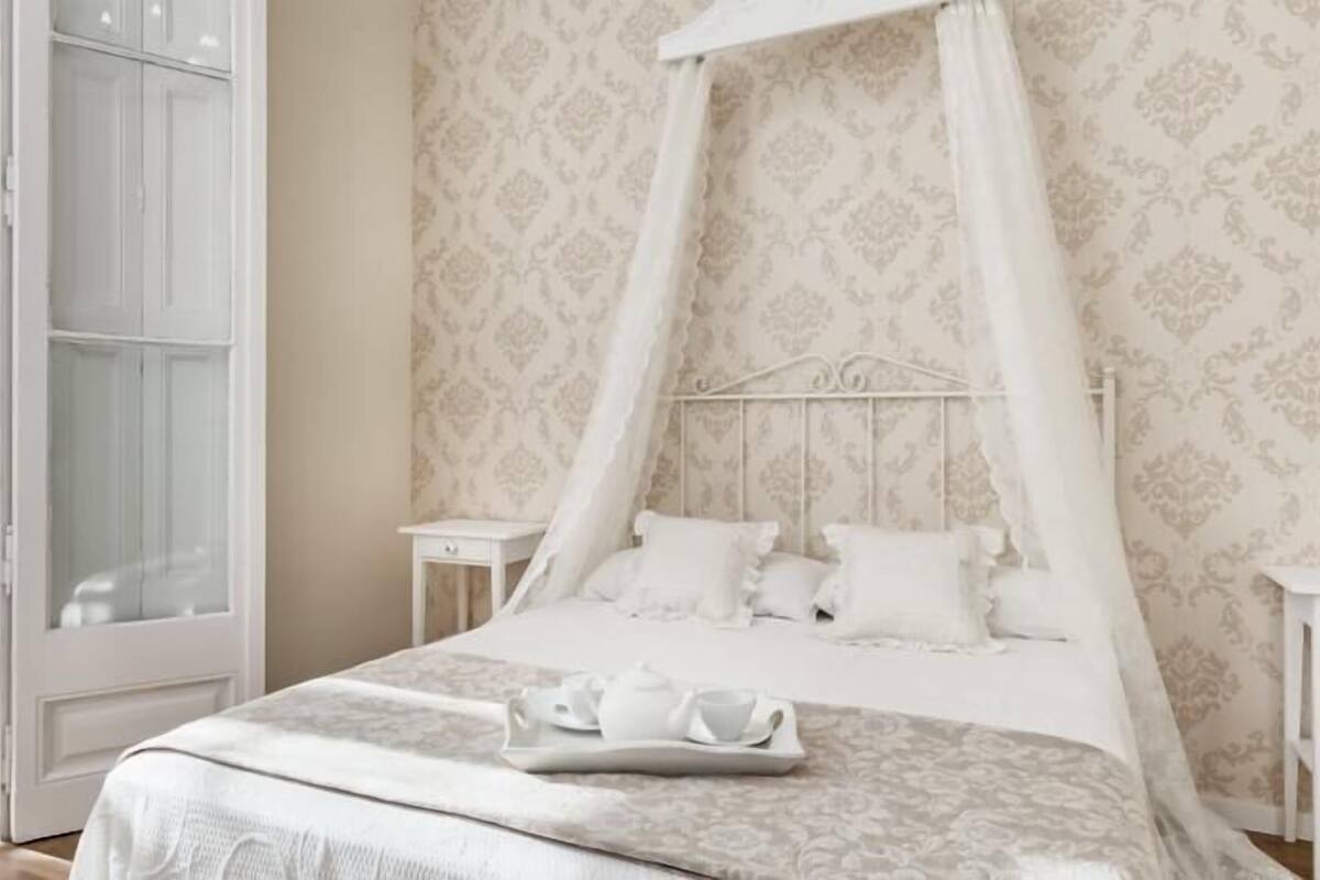 A bedroom with a canopy bed, a bedside table, a nightstand, and a window with a view.
