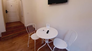 Een kleine kamer met een ronde tafel, twee witte stoelen, een aan de muur bevestigde televisie en een trap naar een andere kamer.