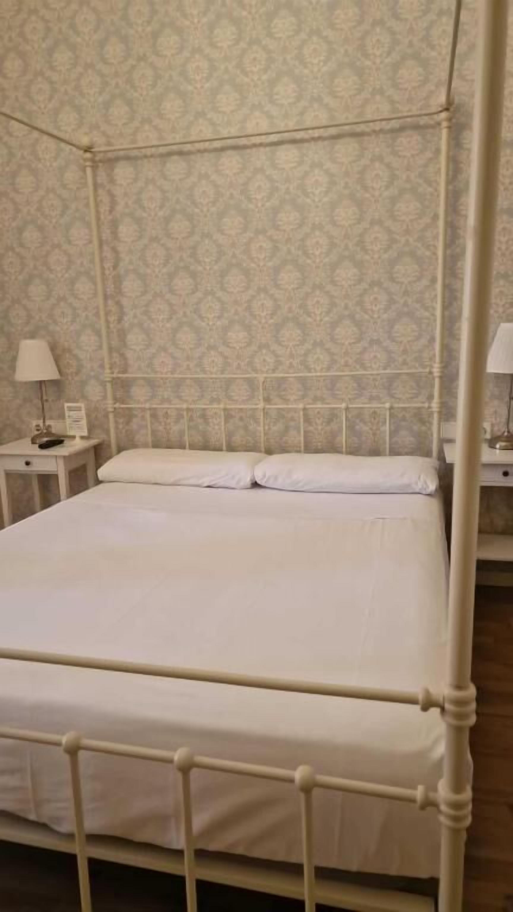 Ein Himmelbett mit weißem Gestell und Bettzeug, ein Nachttisch mit Lampe sowie eine Wand mit ornamentalem Wandpapier.