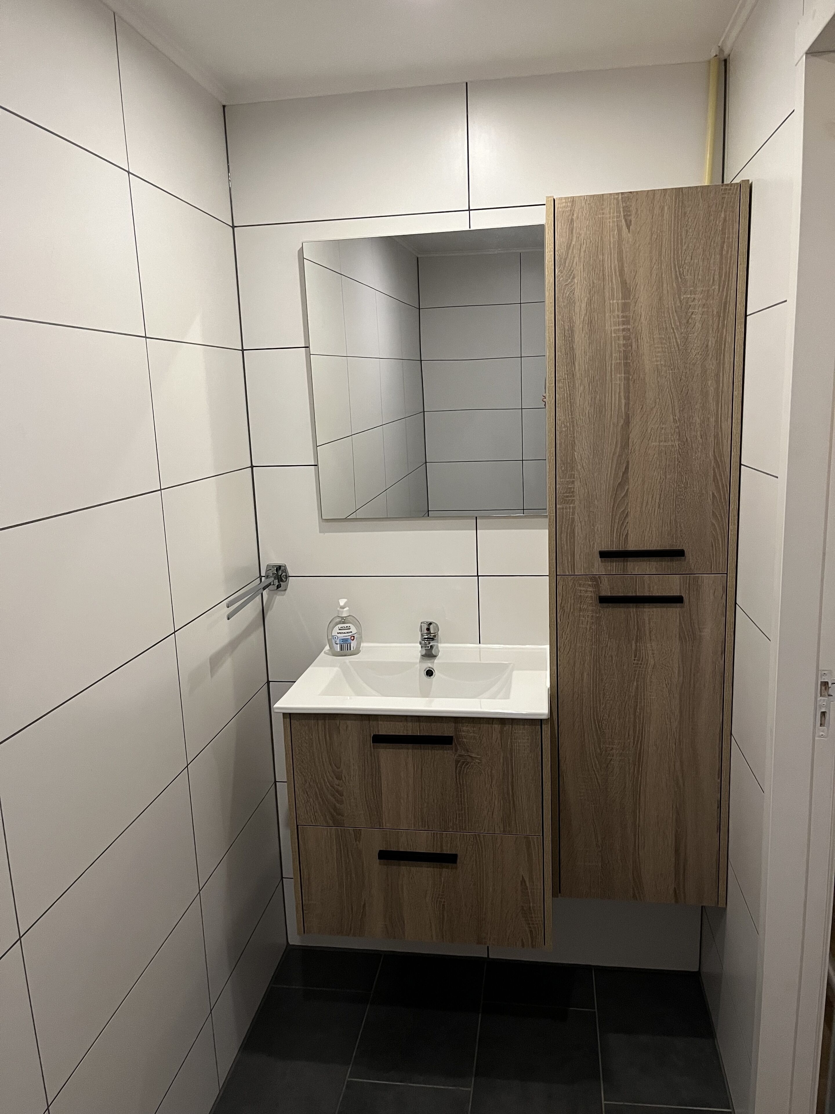 Un baño con paredes de azulejos blancos, un tocador de madera con espejo, un lavabo y piso negro.
