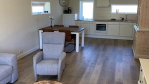 A spacious living area with a dining table, two armchairs, and a coffee table. A kitchen with white cabinets and a window is visible in the background. - Quiet recreation house in Zeeland with Netflix 2 (Bruinisse)