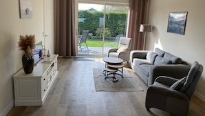 A modern living room with a grey sofa, a small round table, a cabinet, and a sliding glass door leading to a garden. - Quiet recreation house in Zeeland with Netflix 2 (Bruinisse)