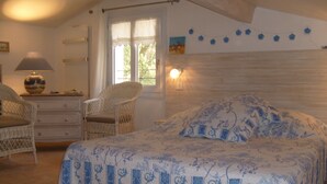 Ein Schlafzimmer mit einem Bett, zwei Korbstühlen, einer Kommode, einer Lampe und einem Fenster mit Vorhängen.