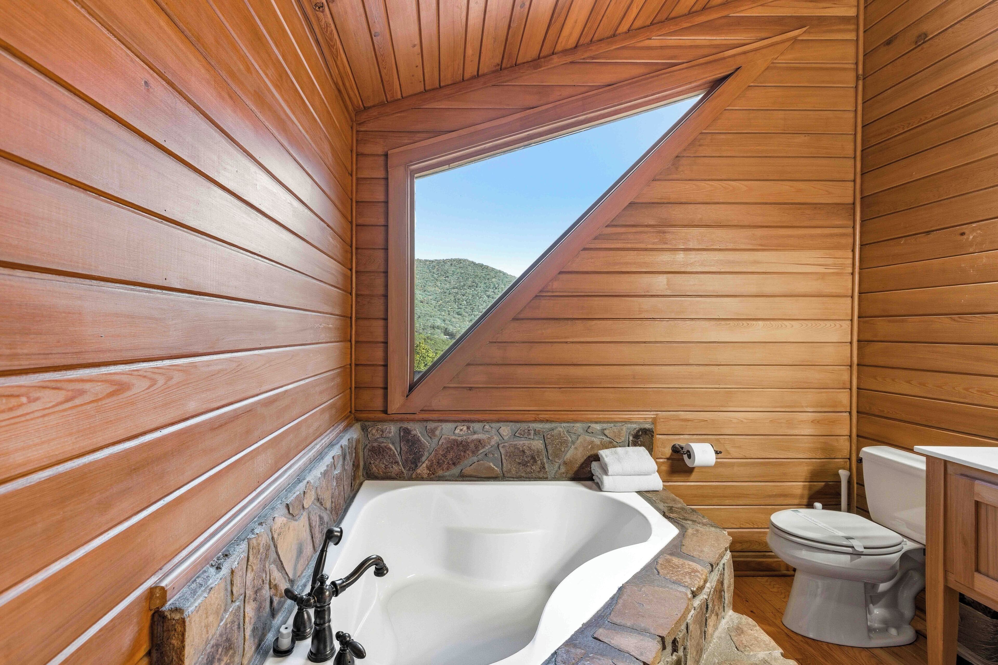A wooden cabin bathroom with a stone-tiled bathtub, a toilet, and a skylight.