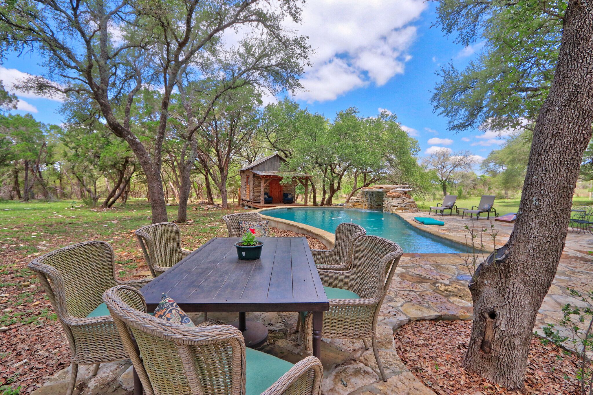 A wooden table with wicker chairs, a small wooden structure, and a swimming pool in a wooded area.