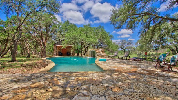 Un patio de piedra con piscina, rodeado de árboles y mobiliario exterior.
