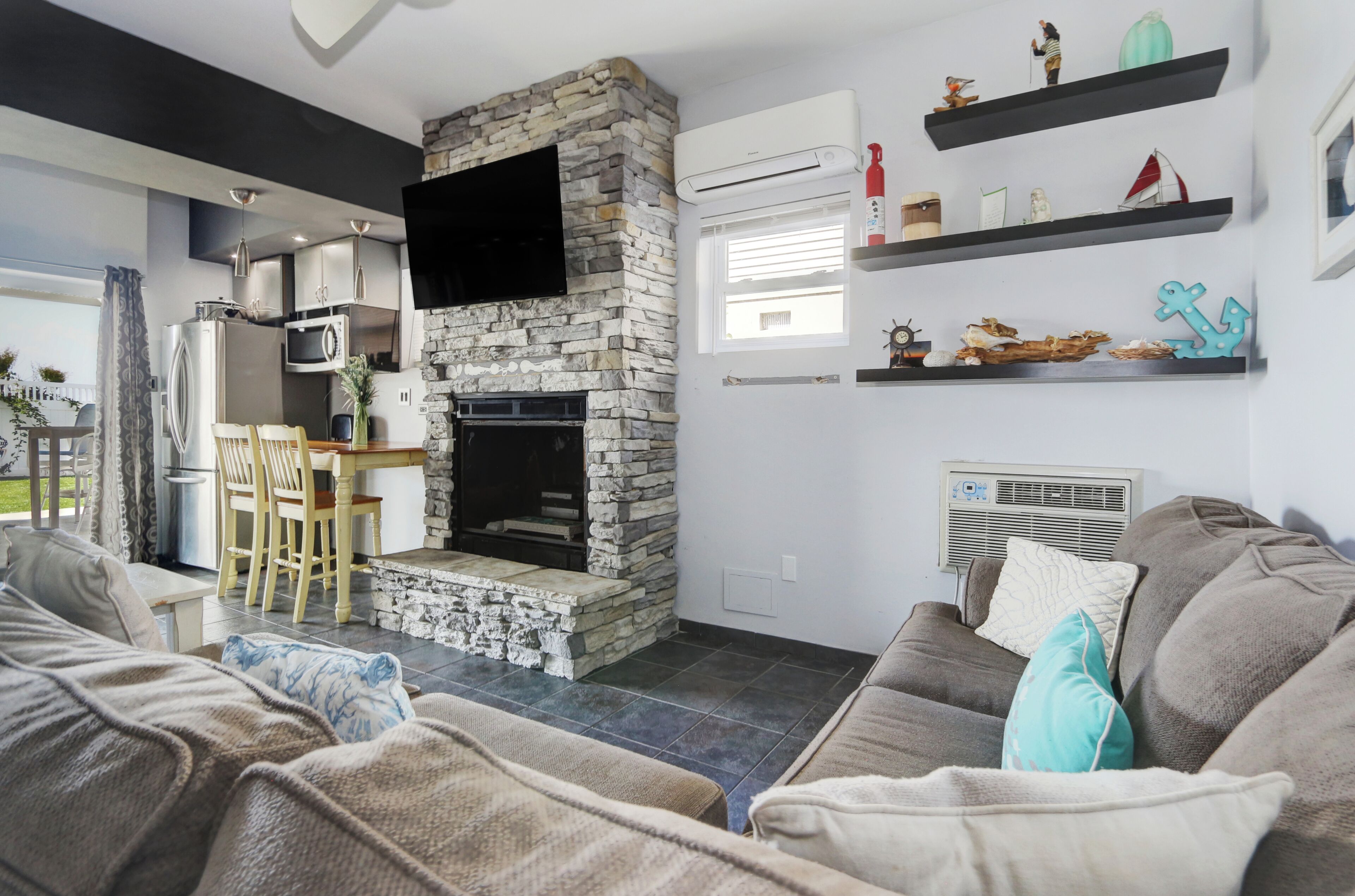 A modern living room with a stone fireplace, a flat-screen TV, a dining area with a table and chairs, and a sofa with cushions.