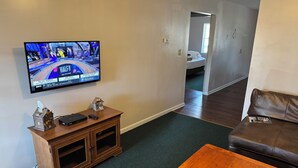 Ein Wohnzimmer mit Fernseher, der ein Sportspiel zeigt, einem Holzschrank, einer braunen Ledersofa und einem Holztisch.