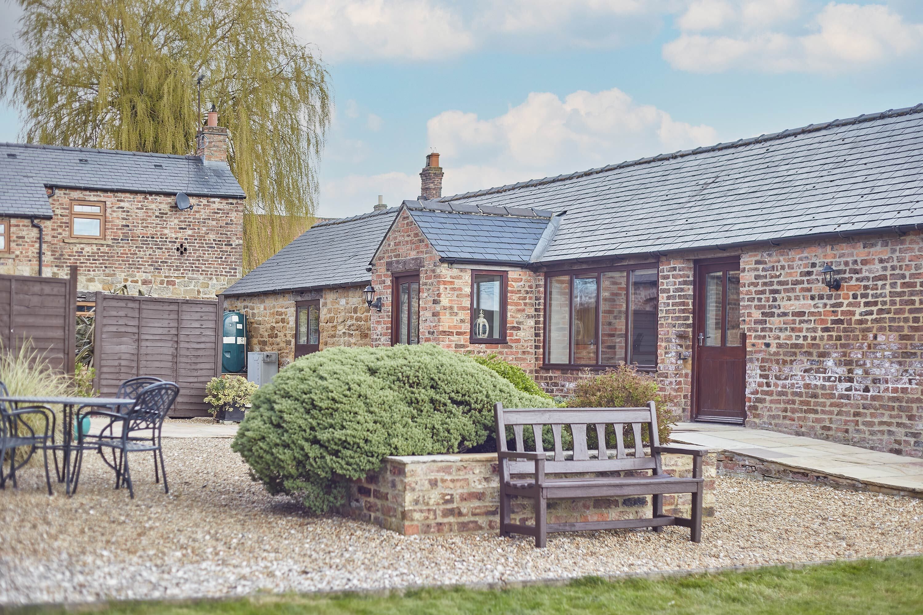 A stone building with a sloped roof, a wooden bench, and a small garden area with a hedge.