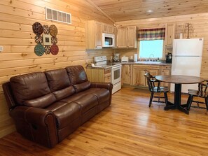 A cozy cabin-style kitchen with wooden walls, a brown leather couch, a dining table with four chairs, and basic kitchen appliances.