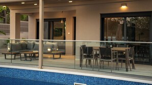 A modern outdoor pool area with a glass railing, a seating area with a sofa and chairs, and a dining table. - Amelia Residence (Zakynthos)