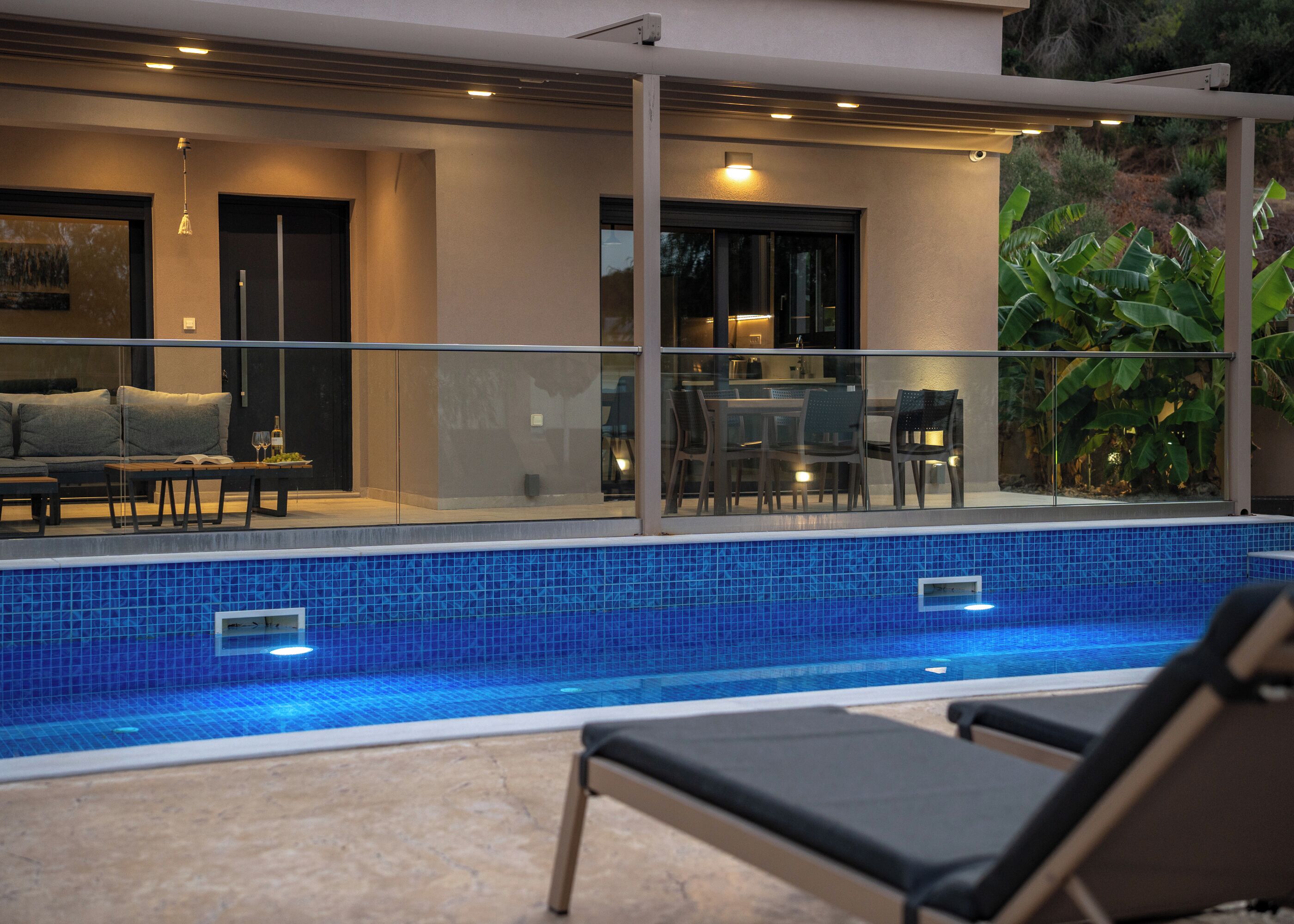 A modern outdoor pool area with a glass balustrade, lounge chairs, and a dining area with a table and chairs.