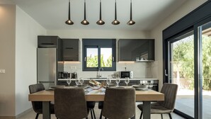 A modern kitchen with a dining area, featuring a large table with chairs, overhead pendant lighting, and a view of greenery outside. - Amelia Residence (Zakynthos)