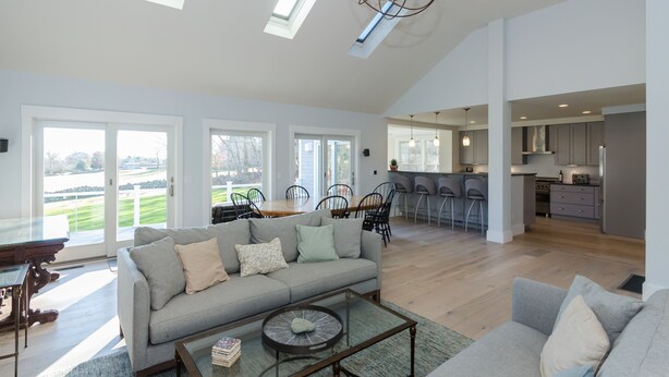 A spacious living area with a grey sofa, a coffee table, and a dining table with chairs. There is a kitchen area with modern appliances and a bar counter. Large windows provide natural light and views of the outdoors. - Costal Country Summer Home with Beach Access - 3 building compound (Westport)