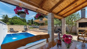 A poolside area with a wooden bench, a table with a wine bottle and glass, and hanging flower baskets. - Nina in Peroj  - Haus für 6 Personen (Fazana)