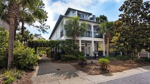 A two-story house with a balcony, surrounded by lush greenery and a paved driveway.