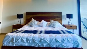 Een moderne slaapkamer met een groot bed, twee nachtkastjes en uitzicht op de zee door een groot raam.