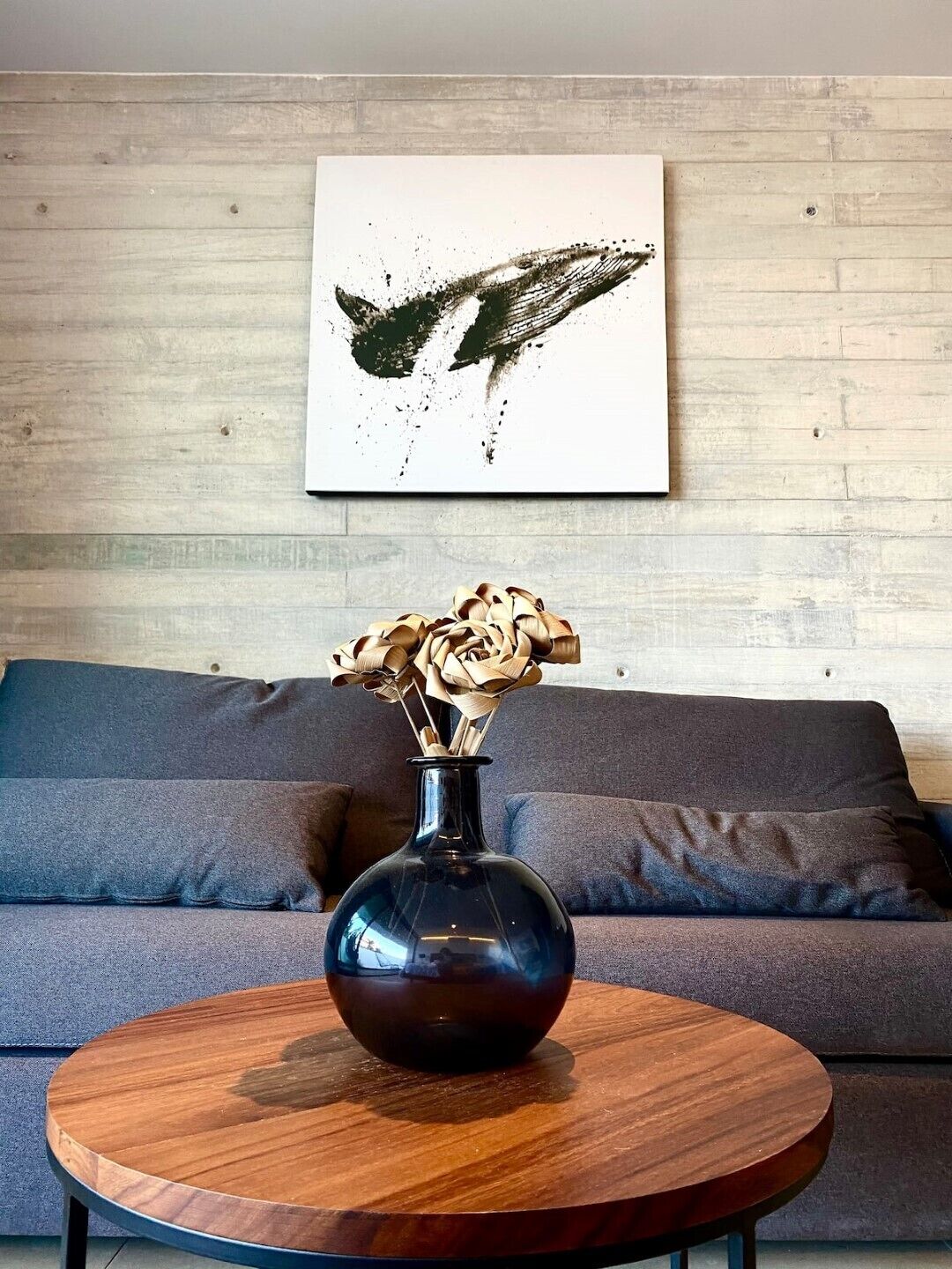 A modern living room with a round wooden coffee table, a dark vase with dried flowers, and a grey sofa. A large abstract painting is on the wall above the sofa.
