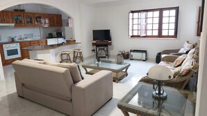A living room with a sofa, a glass coffee table, a television, and a kitchen area with wooden cabinets and appliances.