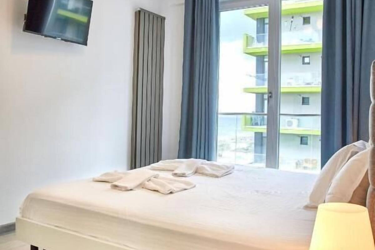 Une chambre d’hôtel avec un lit, un téléviseur fixé au mur, une fenêtre avec des rideaux et une table de chevet avec une lampe.