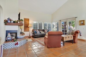 A living room with a fireplace, sofa, armchair, and a window with blinds. - La Hacienda | 3BR Santa Rosa Cove (La Quinta)