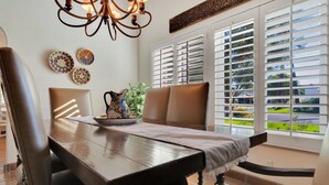 A dining area with a wooden table, chairs, a chandelier, decorative plates, and a view through large windows with blinds.