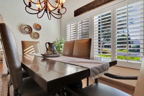 A dining area with a wooden table, chairs, a chandelier, decorative plates, and a view through large windows with blinds.