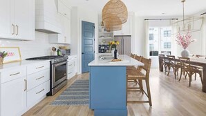 A modern kitchen with white cabinetry, a blue island, and a dining area with wooden chairs. - The Jolene – Rooftop w/ Firepit, Close to 12 South (Nashville)