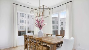 A dining area with a wooden table, white chairs, a vase with flowers, and large windows with a view of buildings. - The Jolene – Rooftop w/ Firepit, Close to 12 South (Nashville)