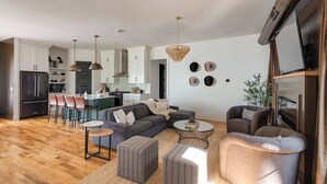 A modern living room with a grey sofa, ottomans, and a coffee table. There is a kitchen in the background with white cabinets and a black refrigerator. - The Gambler - Pool Table + Rooftop Lounge! (Nashville)