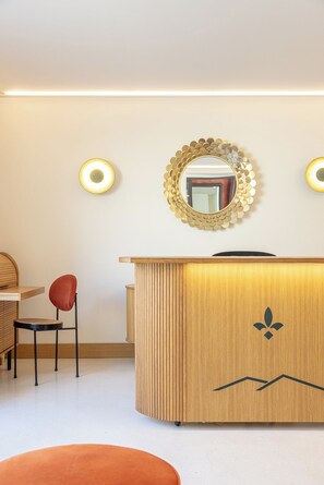 Una recepción con mostrador de madera, un espejo redondo con marco dorado y dos luces fijadas a la pared.