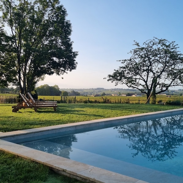 Una piscina con zona verde y árboles, un banco y vistas a campos lejanos.