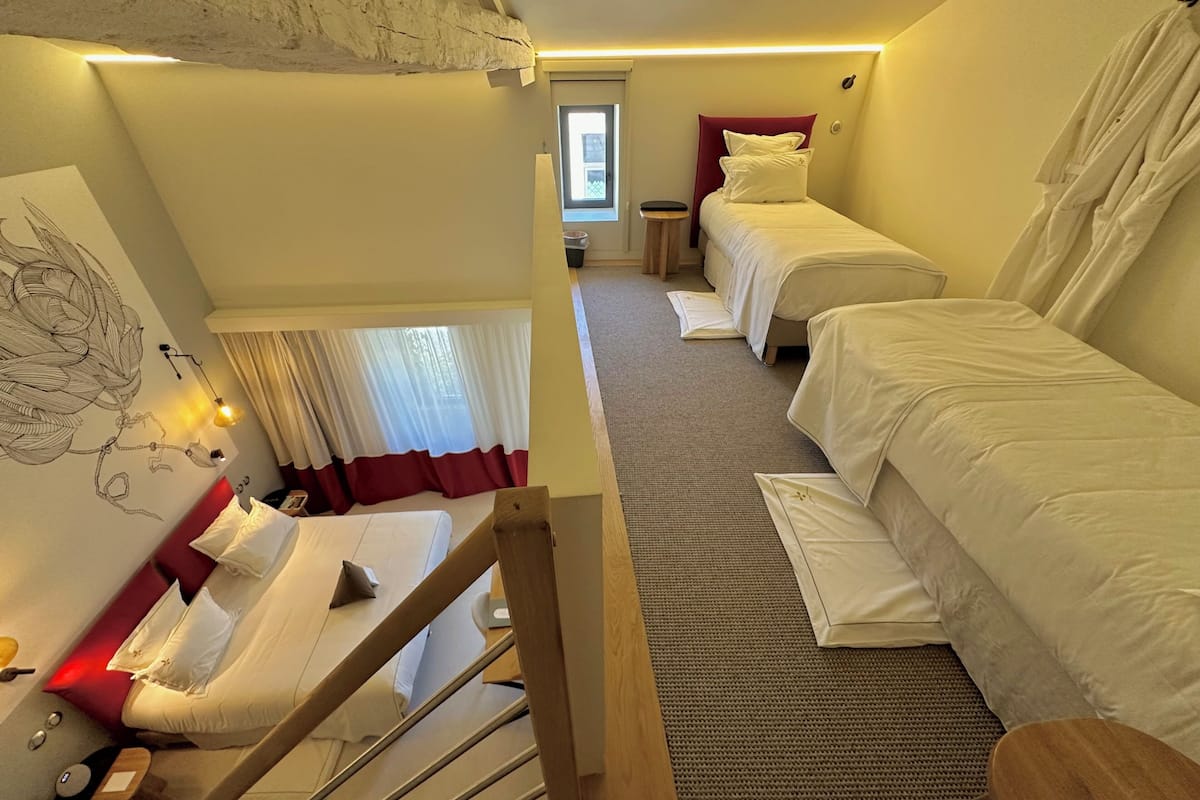 A hotel room with a bunk bed, a small table, a chair, a window with curtains, and a wall art piece.