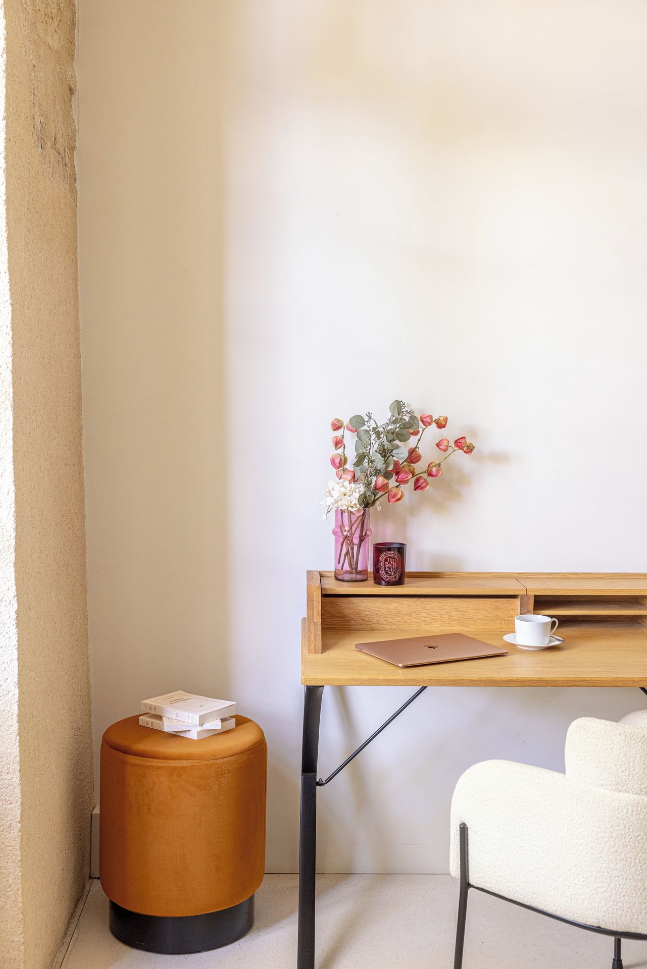 Une petite pièce chaleureuse, avec un bureau en bois, une chaise blanche, un pouf rond et un vase avec des fleurs accroché au mur.