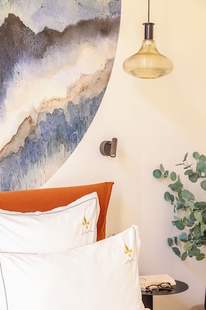 Un dormitorio con cama, una lámpara de pared, una obra de arte en la pared y una mesita con una planta y un libro.