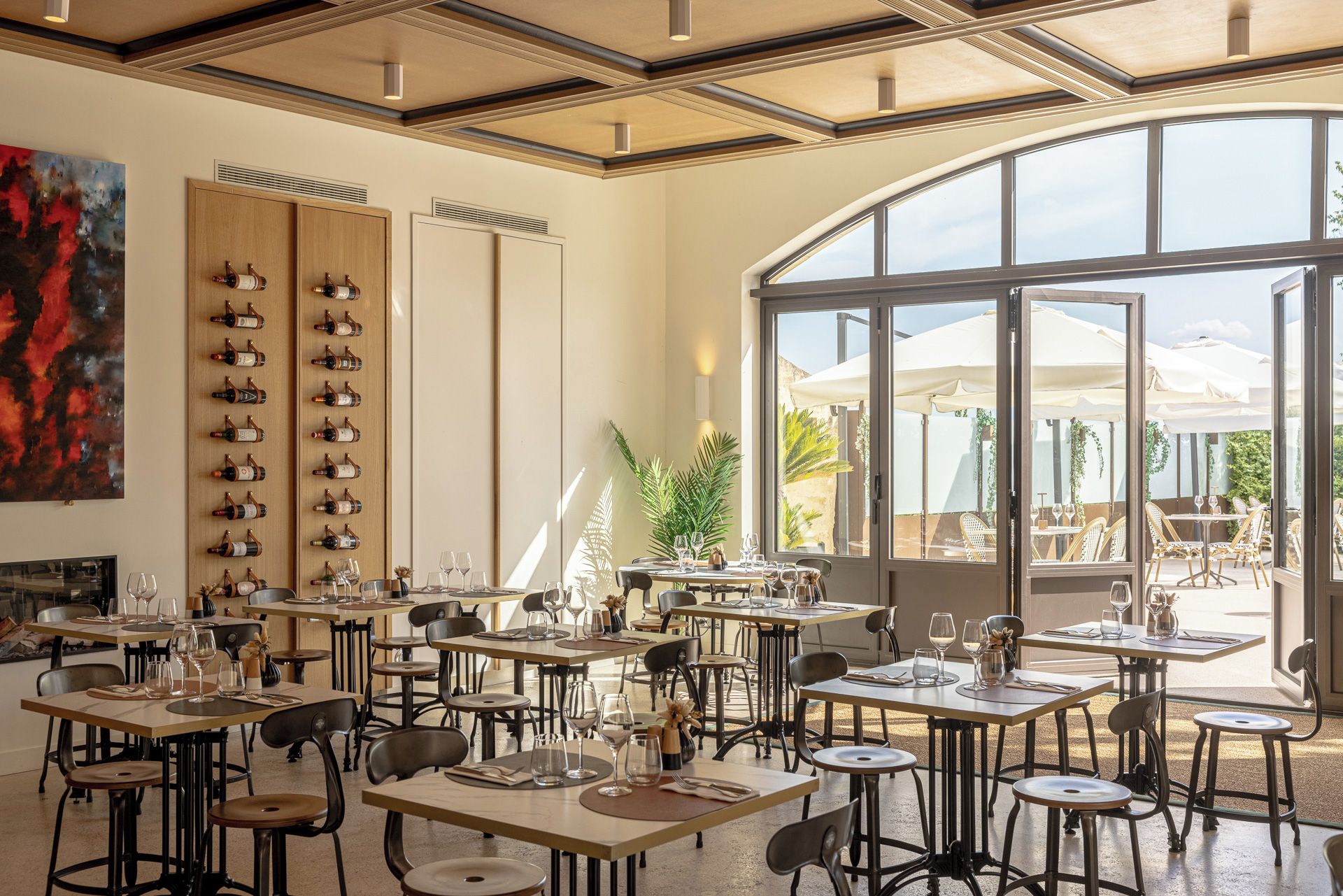 A restaurant with multiple tables and chairs, a wine rack on the wall, and a large window overlooking an outdoor area.