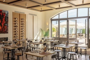Un restaurante con varias mesas y sillas, una estantería para vinos en la pared y un amplio ventanal con vistas a un espacio exterior.