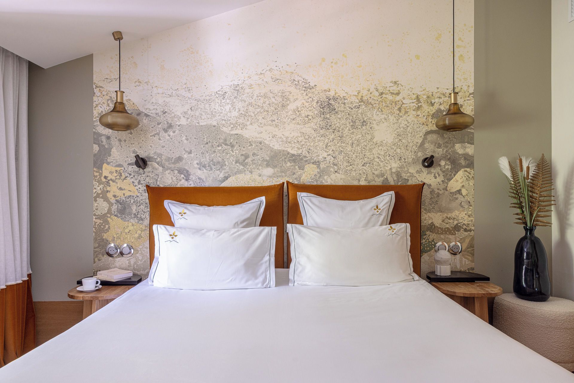 A double bed with white linens and two pillows, a wooden headboard, a nightstand with a lamp, and a vase with dried plants on the right.
