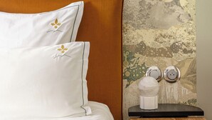 Una cama con sábanas blancas y un cabecero marrón, una mesita de noche redonda de madera con un objeto blanco y una pared con textura que tiene una lámpara colgante.