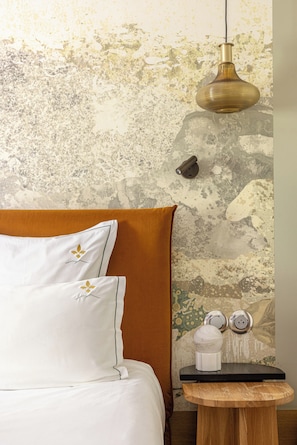 Una cama con sábanas blancas y un cabecero marrón, una mesita de noche redonda de madera con un objeto blanco y una pared con textura que tiene una lámpara colgante.